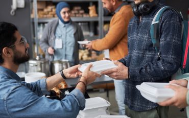 Side view closeup of volunteer giving free food to Middle Eastern refugees standing in line at soup kitchen