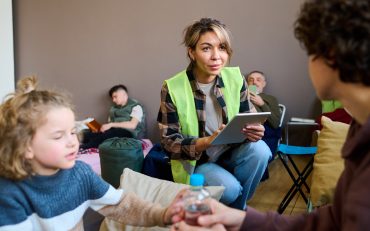 Young female volunteer with tablet sitting on squats in front of woman with son and entering their names and other personal data